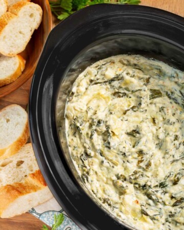 Slow Cooker Spinach Artichoke Dip in black crock with sliced baguette slices around it.