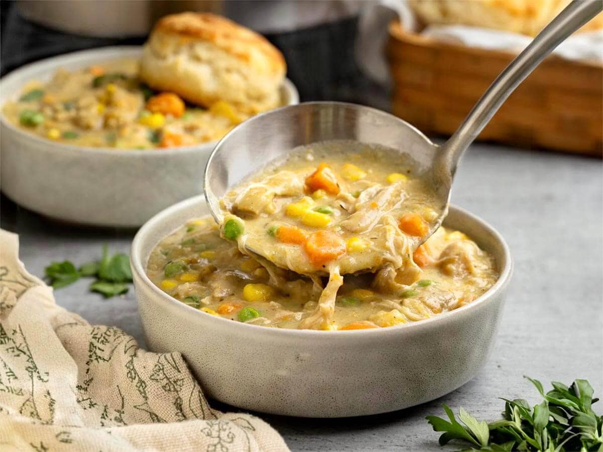 Ladle lifting creamy Instant Pot Chicken Pot Pie filling with shredded chicken, carrots, peas, and corn from a bowl