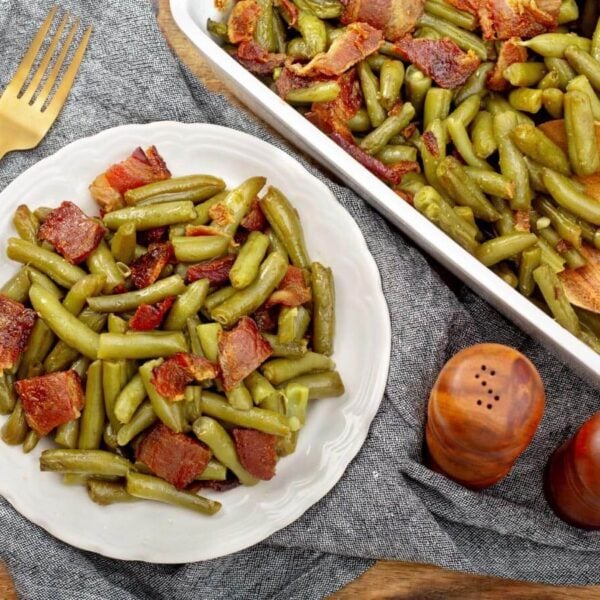 Brown sugar bacon green beans on a white plate beside the baking dish and a wooden serving spoon