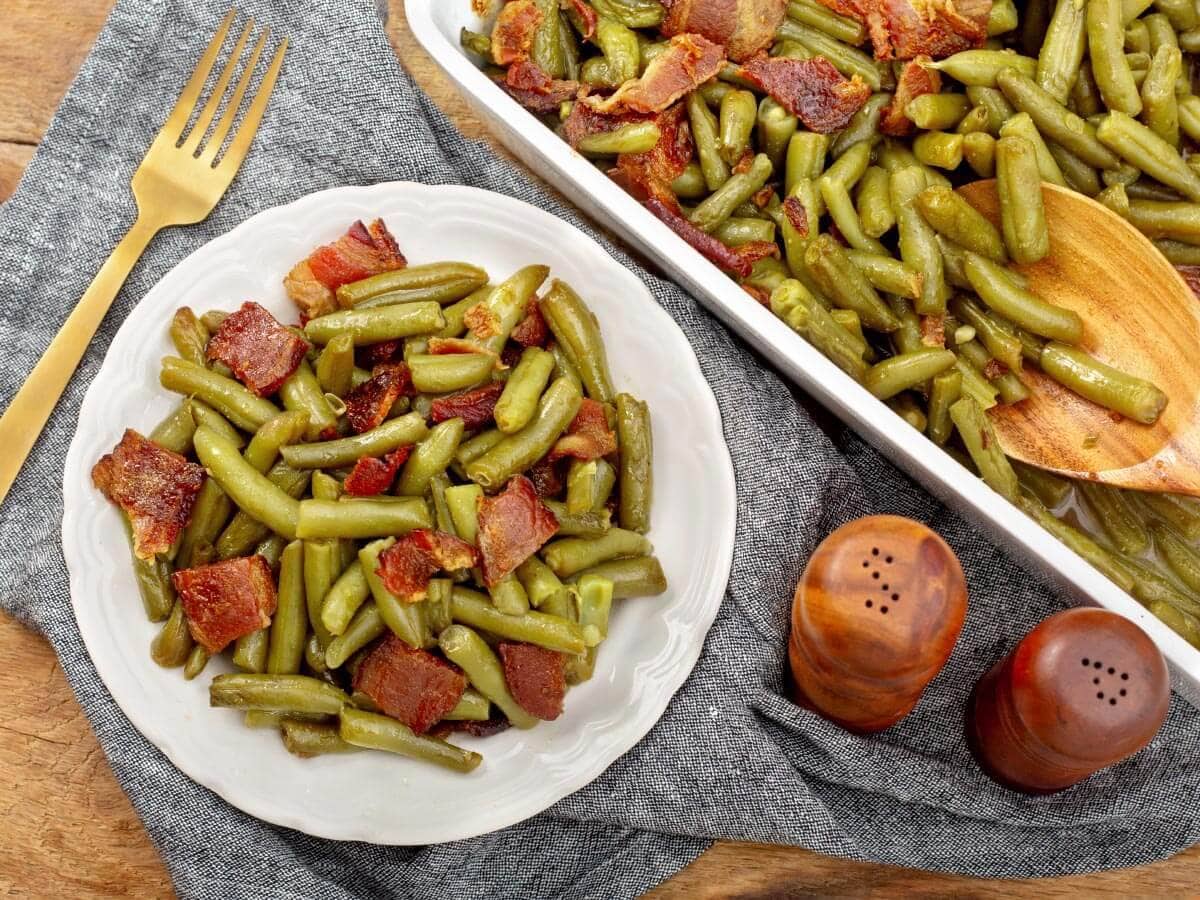 Brown sugar bacon green beans on a white plate beside the baking dish and a wooden serving spoon
