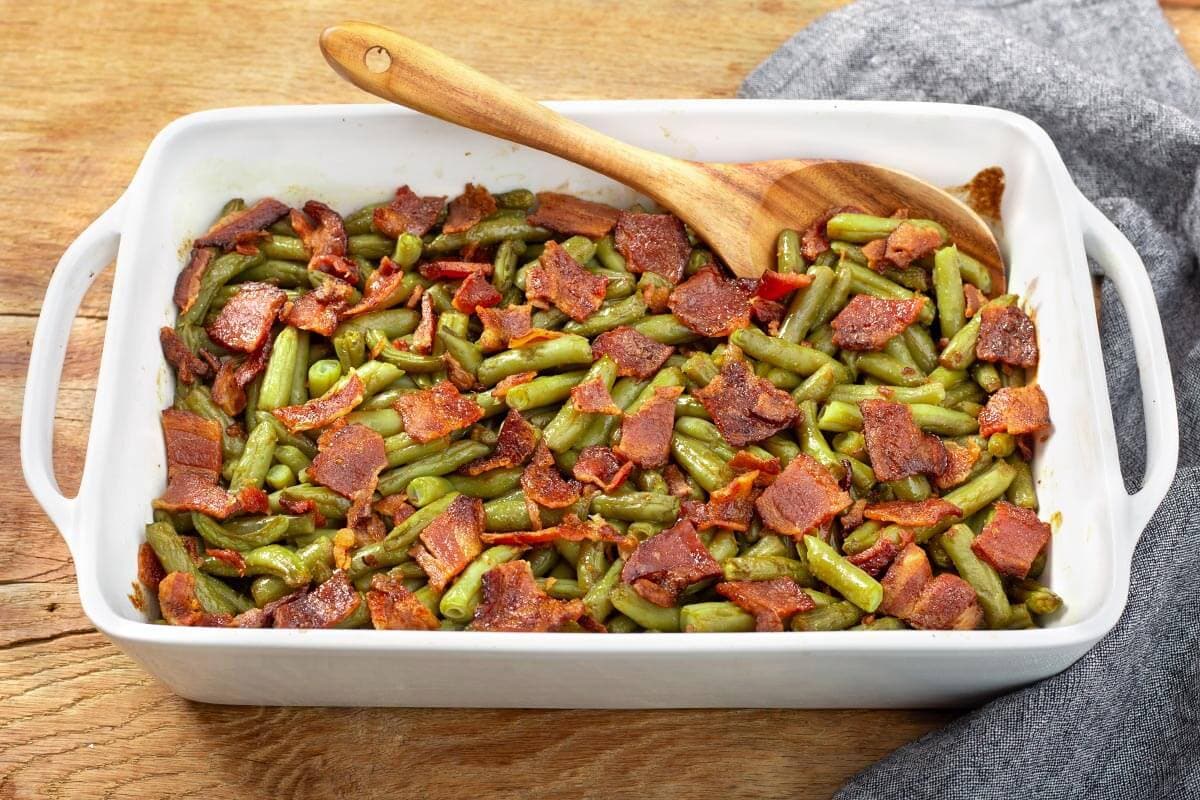 Baked green beans topped with chopped bacon in a white casserole dish with a wooden serving spoon