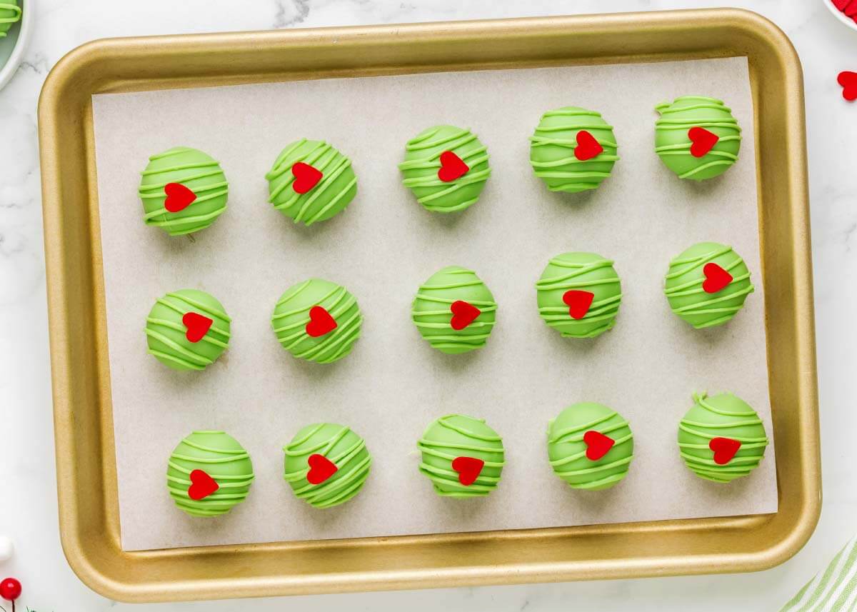 Grinch Truffles decorated with green candy drizzle and red hearts on a parchment-lined baking sheet
