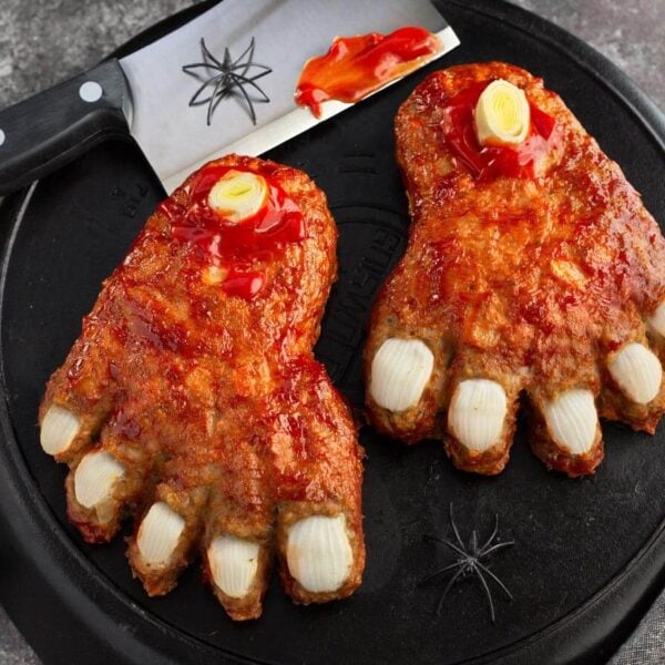 Halloween Feet Loaf with onion toenails, leek bones, and ketchup glaze on black cast iron beside a cleaver and spiders.