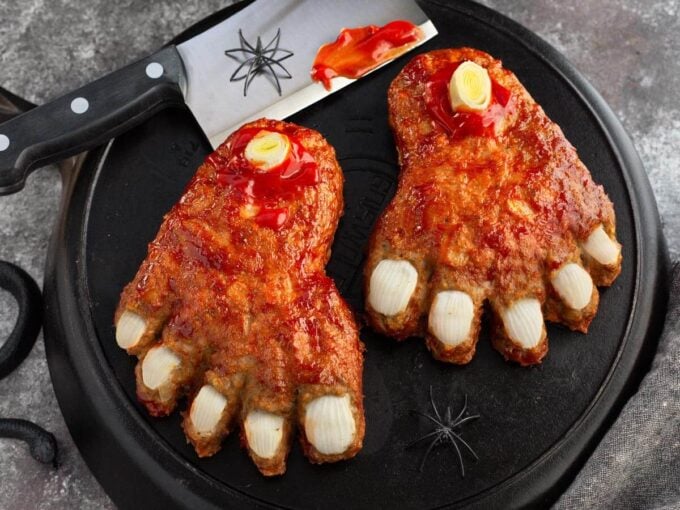 Halloween Feet Loaf with onion toenails, leek bones, and ketchup glaze on black cast iron beside a cleaver and spiders.