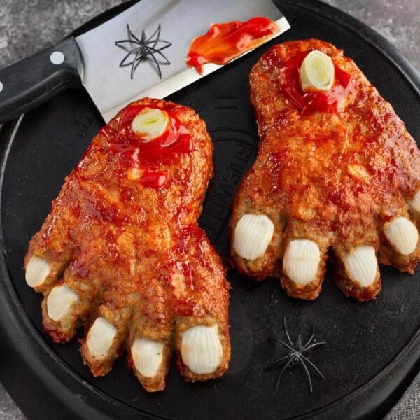 Halloween Feet Loaf with onion toenails, leek bones, and ketchup glaze on black cast iron beside a cleaver and spiders.