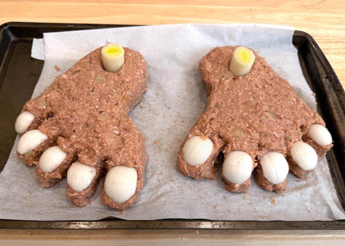 Two uncooked ground turkey meatloaf feet with onion toenails and leek ankle bones on a parchment-lined baking sheet.