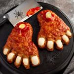 Two glazed Halloween meatloaf feet with onion toenails and leek bones on black cast iron with a cleaver and plastic spiders.