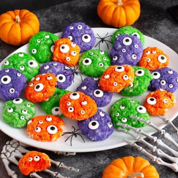 Green, purple, and orange Halloween Monster Cookies with candy eyes on a white platter surrounded by pumpkins and spooky decorations.
