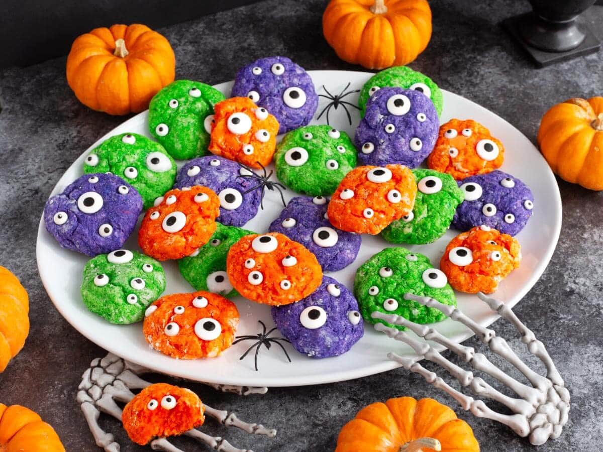 Green, purple, and orange Halloween Monster Cookies with candy eyes on a white platter surrounded by pumpkins and spooky decorations.