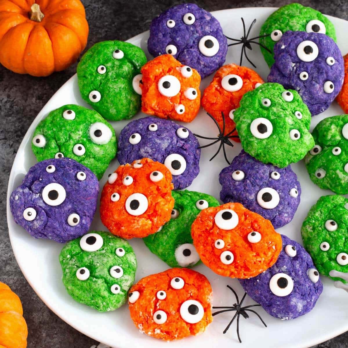 Close-up of orange, green, and purple Halloween Monster Cookies with googly candy eyes and spider decorations on a white plate.