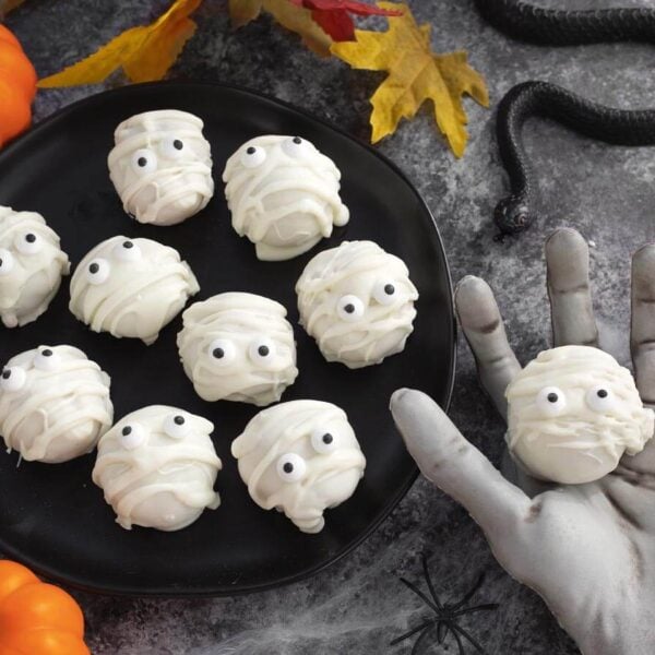 White chocolate mummy Oreo truffles with candy eyes on a black plate beside Halloween decorations