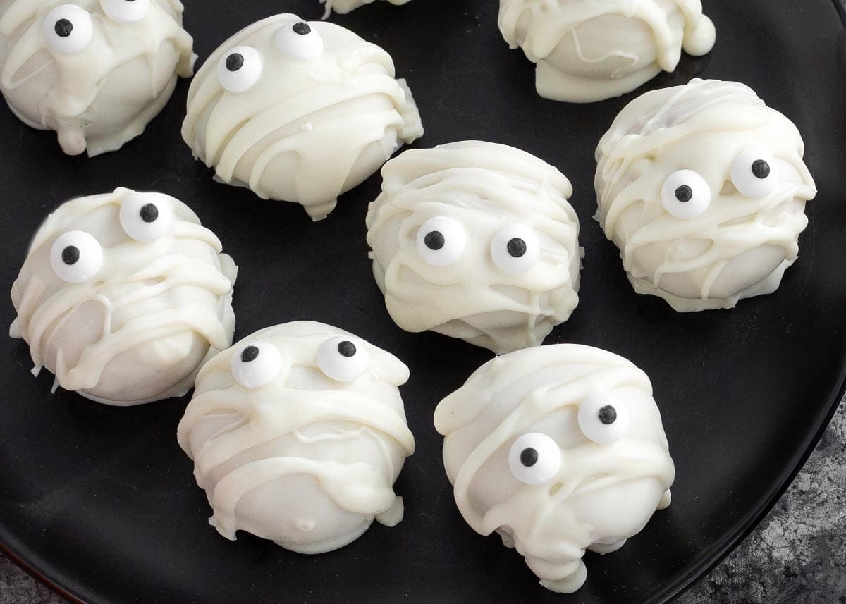 Close-up of mummy Oreo truffles with white chocolate bandages and candy eyes on a black plate