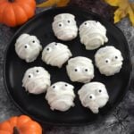 White chocolate mummy Oreo truffles with candy eyes on a black plate surrounded by pumpkins and Halloween decorations
