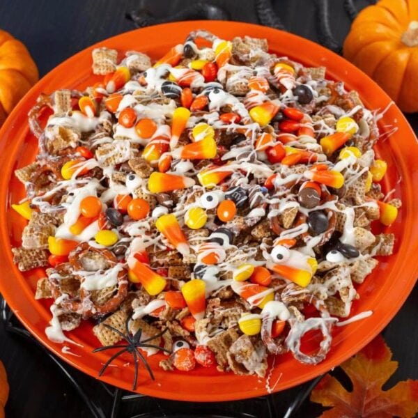 Halloween Snack Mix with Cinnamon Chex, pretzels, white chocolate drizzle, and colorful Halloween candy on an orange platter with pumpkins.