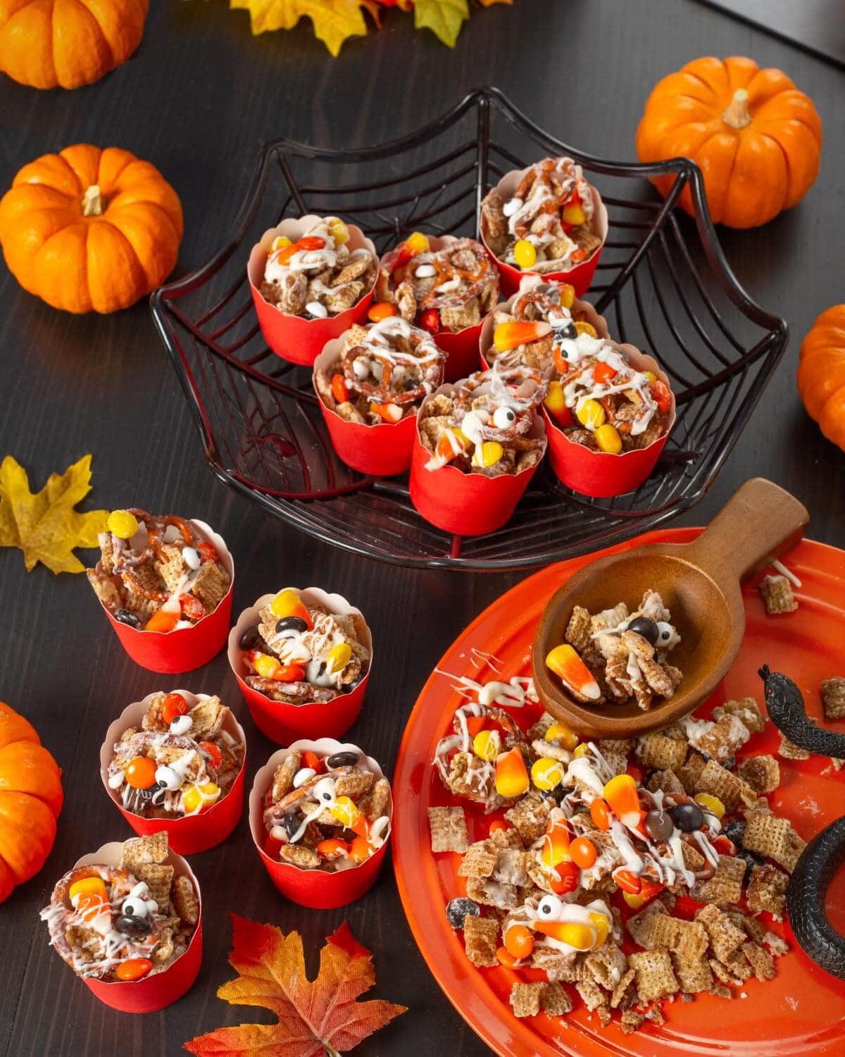 Individual cups of Halloween Snack Mix arranged in a black spiderweb basket, with more snack mix on an orange platter.