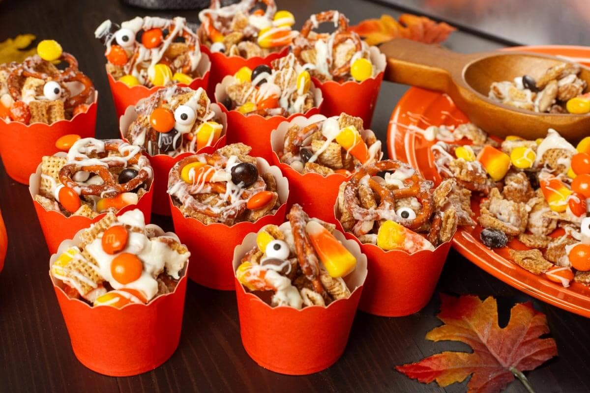 Halloween Snack Mix served in red scalloped paper treat cups with candy corn, chocolate candies, and white chocolate drizzle.