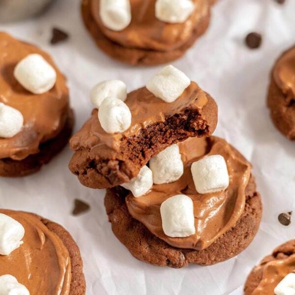 Hot cocoa cookies with chocolate frosting and mini marshmallows, with a bite taken out of one to show the chocolate center