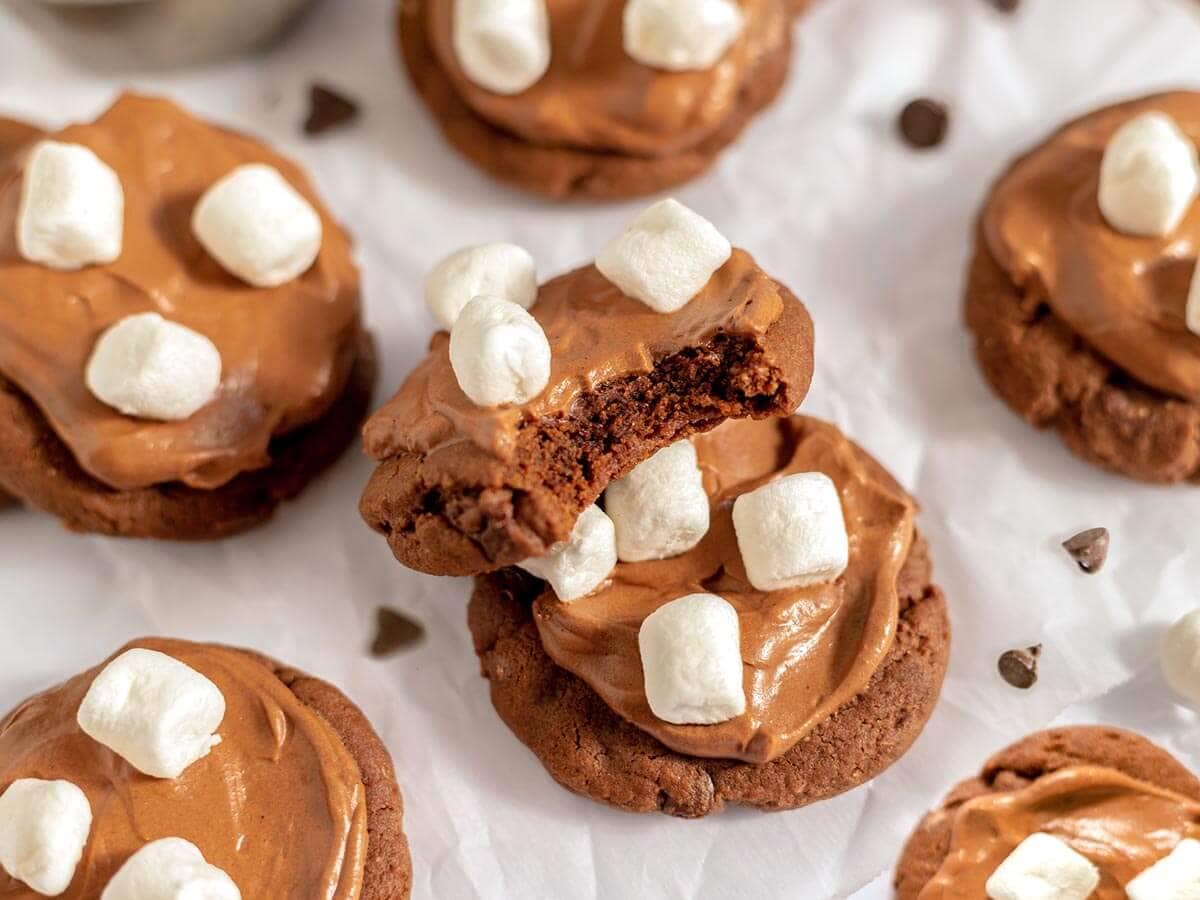 Hot cocoa cookies with chocolate frosting and mini marshmallows, with a bite taken out of one to show the chocolate center