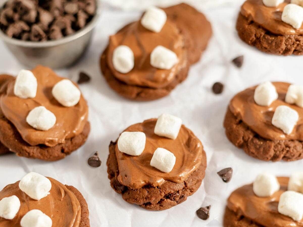 Hot cocoa cookies topped with swirls of chocolate frosting and mini marshmallows, arranged on parchment paper