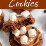 Hot cocoa cookies with chocolate frosting and mini marshmallows, with a bite taken out of one to show the chocolate center