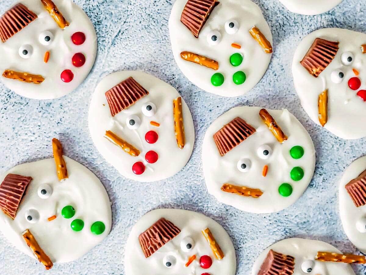 Melted snowman bark with peanut butter cup hats, candy eyes, red and green buttons, and pretzel arms.