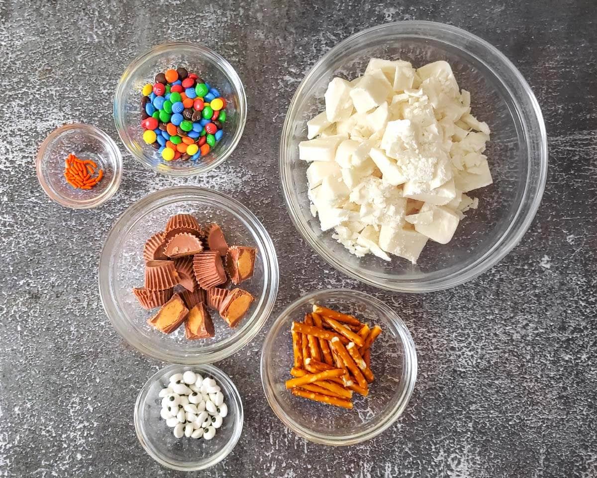 Bowls of chopped white almond bark, peanut butter cup halves, mini M&M’s, candy eyes, orange sprinkles, and pretzel pieces.