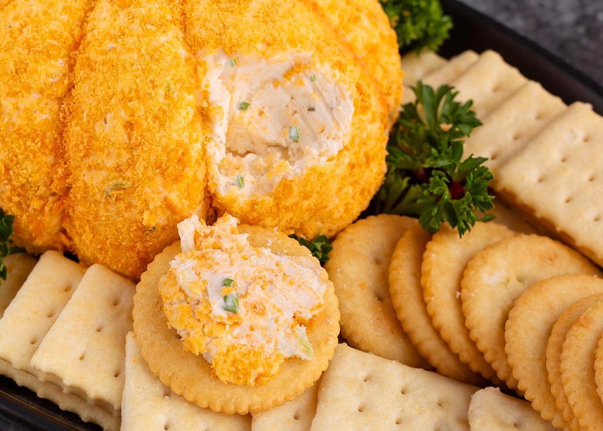 Pumpkin cheese ball closeup with a scoop removed, showing the creamy cheddar filling and a cracker topped with cheese spread.