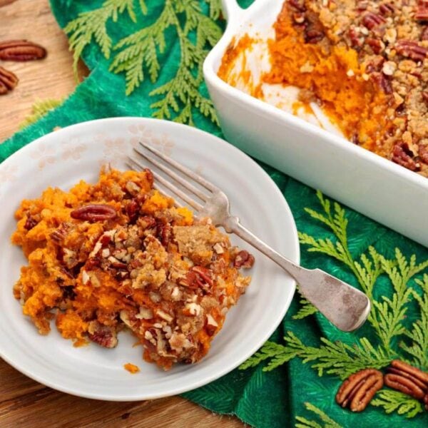 Sweet potato casserole with pecan topping on a white plate beside the baking dish, with pecans scattered on the wooden table