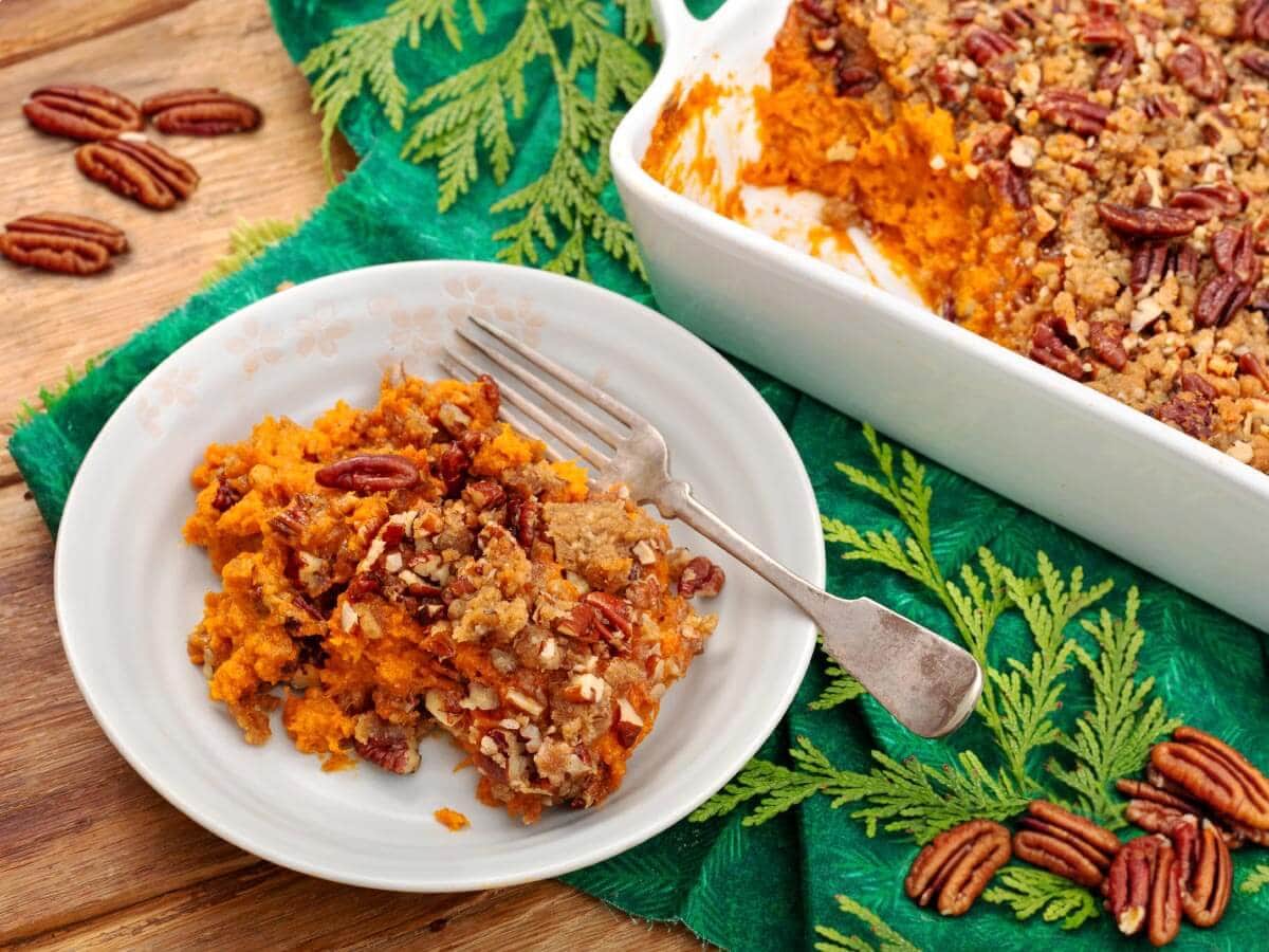 Sweet potato casserole with pecan topping on a white plate beside the baking dish, with pecans scattered on the wooden table
