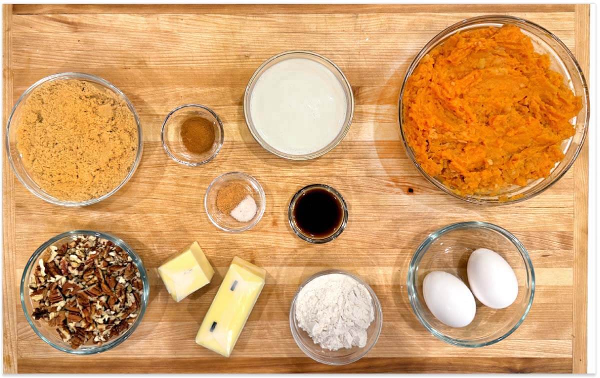 Bowls of mashed sweet potatoes, brown sugar, milk, spices, vanilla, pecans, and flour with butter and eggs on a wooden board