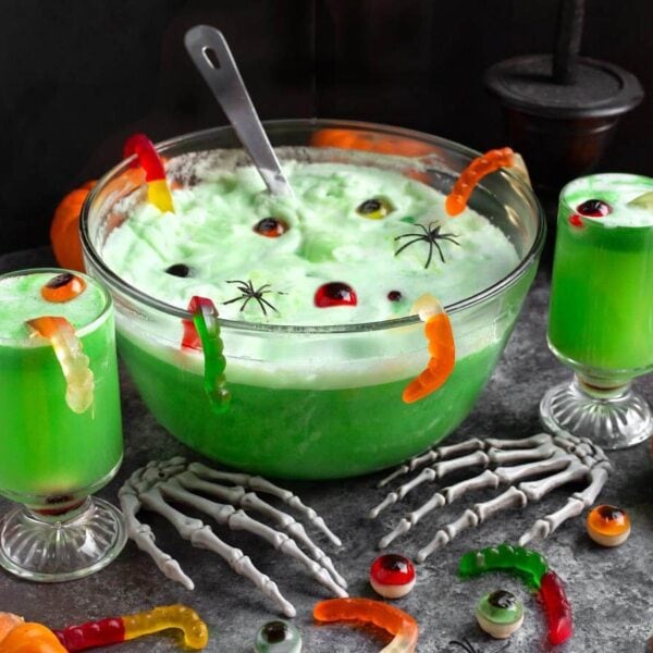 Green Witches Brew Halloween Punch in a glass punch bowl with two serving glasses, pumpkins, and spooky Halloween decorations.