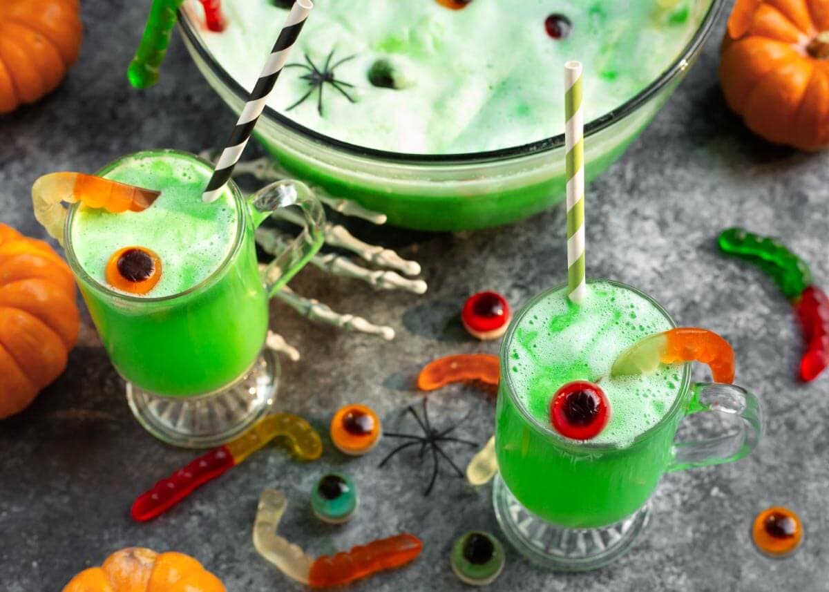 Two glasses of bright green Halloween punch with candy eyeballs and gummy worms beside a frothy punch bowl