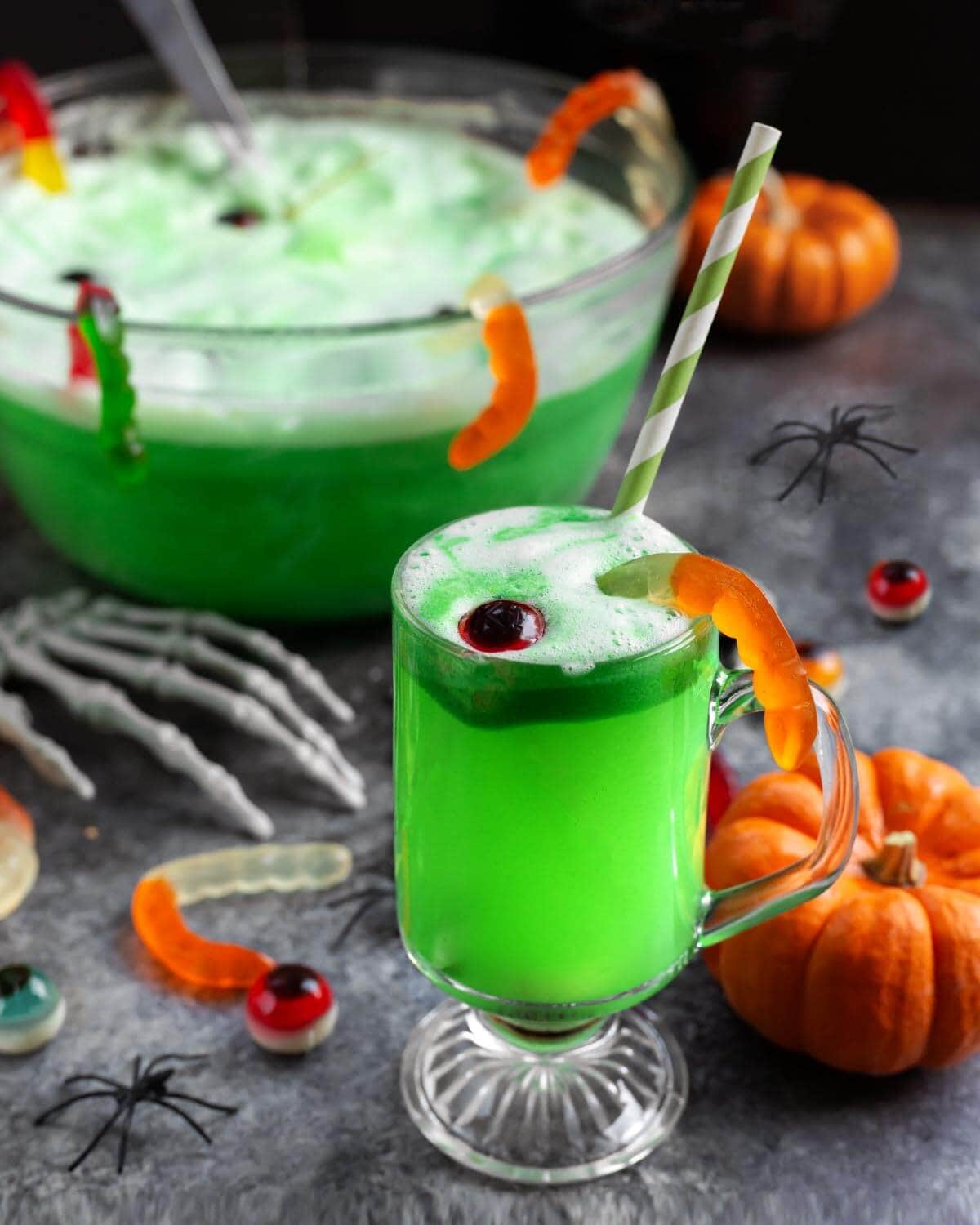 Close-up of a glass of green Halloween punch decorated with a candy eyeball and gummy worm, with a punch bowl in the background.