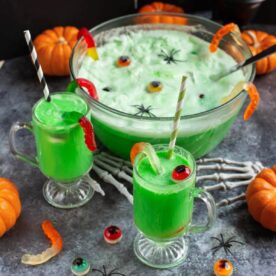 Witches Brew Halloween Punch with bubbly green lime sherbet foam, served in a glass punch bowl with two decorated drinks.